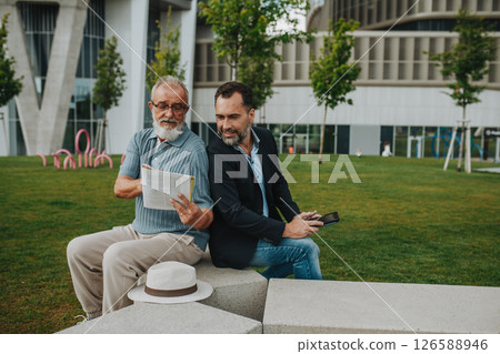 Father and adult son sitting on bench in city park, reading newspaper. 126588946