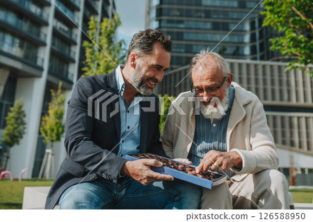 Senior dad and adult son sharing a box of sweets, chocolates on park bench. Senior dad and adult son sharing a box of sweets, chocolates on park bench. 126588950