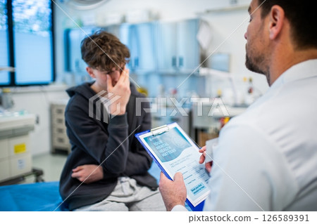 Depressed teenage patient sitting on hospital bed, talking with caring doctor. 126589391