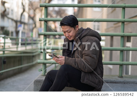 Young Asian man sitting on street ledge scrolling on smartphone 126589542