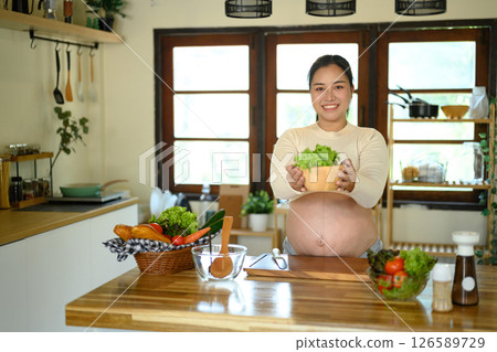 Asian pregnant woman standing in a bright kitchen, presenting a bowl of salad 126589729