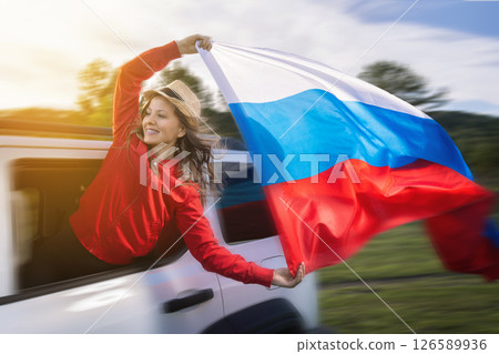 A woman joyfully waves the Russian flag from the window of a moving car as she embarks on a road trip to celebrate Russia Day. Dressed in a red outfit, she embodies the spirit of independence and A woman joyfully waves the Russian flag from the window of a moving car as she embarks on a road trip to celebrate Russia Day. Dressed in a red outfit, she embodies the spirit of independence and 126589936