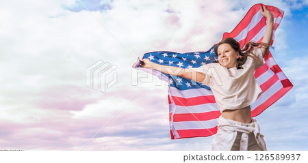 Young American woman stands outdoors, joyfully waving a large American flag usa as celebrates Independence Day outdoors. Her hair blows in the breeze, and her face beams with excitement and pride. Young American woman stands outdoors, joyfully waving a large American flag usa as celebrates Independence Day outdoors. Her hair blows in the breeze, and her face beams with excitement and pride. 126589937