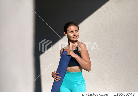 A young woman with a fit physique stands outdoors, holding a yoga mat. Girl looks relaxed and happy, embodying a healthy lifestyle and preparing for her next fitness session A young woman with a fit physique stands outdoors, holding a yoga mat. Girl looks relaxed and happy, embodying a healthy lifestyle and preparing for her next fitness session 126590748