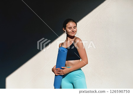 A young woman stands confidently with a blue yoga mat, enjoying the warmth of the sun. Dressed in stylish fitness attire, she embodies health and relaxation during her outdoor activity A young woman stands confidently with a blue yoga mat, enjoying the warmth of the sun. Dressed in stylish fitness attire, she embodies health and relaxation during her outdoor activity 126590750