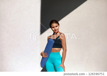 A young, fit woman stands confidently while holding a yoga mat, embracing the outdoors for her exercise routine. Girl radiates happiness and health in a vibrant atmosphere. A young, fit woman stands confidently while holding a yoga mat, embracing the outdoors for her exercise routine. Girl radiates happiness and health in a vibrant atmosphere. 126590751