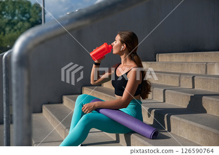 Young athletic woman sitting on concrete steps, enjoying a refreshing drink from a red bottle, with yoga mat beside her, showcasing a healthy lifestyle and fitness routine in a bright outdoor setting 126590764