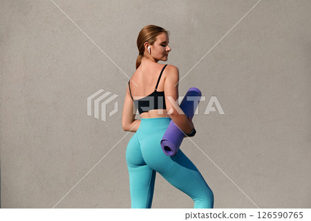 Young woman in athletic wear holding a yoga mat, standing against a neutral background, showcasing fitness lifestyle and dedication to health and wellness, ready for a yoga session or workout 126590765