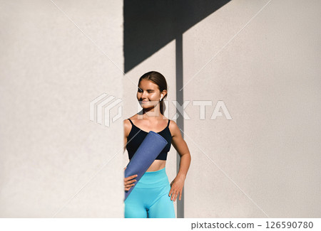 A young woman stands gracefully with a yoga mat, exuding confidence in workout attire. Girl is in an outdoor setting, savoring the beauty of nature while engaging in a healthy lifestyle 126590780