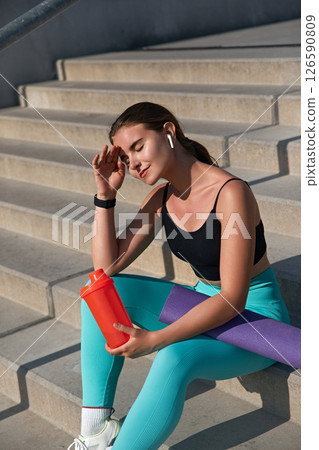 Young athletic woman in stylish activewear sitting on outdoor steps, holding a red shaker bottle, taking a break after workout, enjoying the sun, showcasing fitness lifestyle and healthy living 126590809