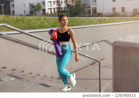 Young woman in athletic wear carrying yoga mat and water bottle, walking down outdoor stairs, surrounded by modern architecture and greenery, embodying a healthy lifestyle and fitness journey 126590813