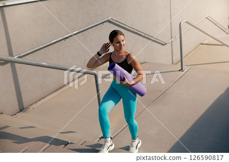 Young athletic woman carrying yoga mat while climbing outdoor stairs, dressed in vibrant activewear, showcasing fitness lifestyle and dedication to health and wellness in a modern urban environment Young athletic woman carrying yoga mat while climbing outdoor stairs, dressed in vibrant activewear, showcasing fitness lifestyle and dedication to health and wellness in a modern urban environment 126590817