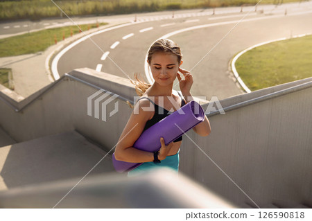Young woman carrying a purple yoga mat while walking up outdoor stairs, dressed in athletic wear, with a sunny environment and winding pathway in the background, promoting fitness and wellness Young woman carrying a purple yoga mat while walking up outdoor stairs, dressed in athletic wear, with a sunny environment and winding pathway in the background, promoting fitness and wellness 126590818