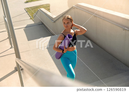 Young woman in athletic wear carrying yoga mat while walking up outdoor stairs, showcasing fitness lifestyle and determination, with bright sunlight illuminating the scene and enhancing the atmosphere 126590819