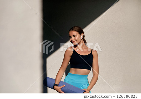 Young woman in athletic wear holding yoga mat, smiling confidently against a minimalist background with soft shadows, embodying a healthy lifestyle and fitness enthusiasm in a serene environment Young woman in athletic wear holding yoga mat, smiling confidently against a minimalist background with soft shadows, embodying a healthy lifestyle and fitness enthusiasm in a serene environment 126590825