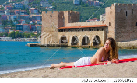 A serene daytime moment unfolds as a person enjoys sunbathing on a sandy beach. The backdrop features an ancient castle with gentle waves lapping at the shore. 126591983