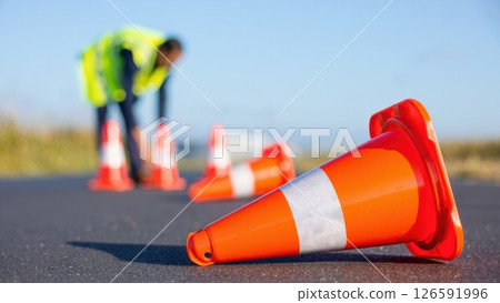 A construction worker adjusts bright orange safety cones on a paved street under clear blue skies. The focus is on the worker's diligence in ensuring safety during roadworks. 126591996