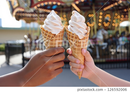 Two joyful hands hold soft serve ice cream cones creating a delightful moment amid the vibrant atmosphere of an amusement park during the sunny afternoon. 126592006
