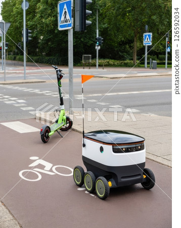 A delivery robot maneuvers through a bike lane while an electric scooter waits nearby. This bustling urban environment showcases modern transportation and efficient technology. 126592034