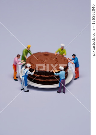 Six miniature bakers in colorful outfits collaborate to lift a sumptuous chocolate cake on a white plate displaying teamwork and culinary delight in a whimsical setting. Six miniature bakers in colorful outfits collaborate to lift a sumptuous chocolate cake on a white plate displaying teamwork and culinary delight in a whimsical setting. 126592040