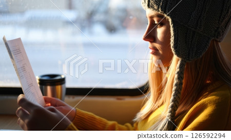 A young woman in a warm hat enjoys a quiet afternoon ride immersed in reading a menu as snow blankets the outside world. The sunlight casts a soft glow on her face adding warmth. 126592294