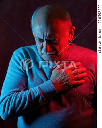A senior man clutches his chest in a moment of distress illuminated by striking red and blue lights. The atmosphere conveys tension and evokes a sense of urgency in his expression. 126592321
