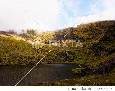 Lough callee landscape displaying calm waters surrounded by vibrant green mountains under a partly cloudy sky in County Kerry, Ireland. 126592487