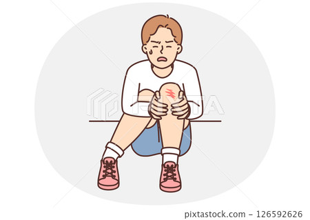 Crying boy sits on ground and holds on to injury on knee after fall during walk 126592626