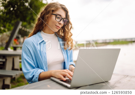 Portrait of woman in blue shirt with laptop at table in outdoor cafe near a lake. Freelance concept. 126593850