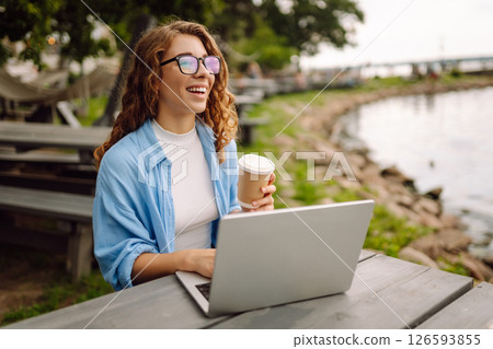 Portrait of a young woman in a blue shirt with a laptop at a table in an outdoor cafe near a lake. Portrait of a young woman in a blue shirt with a laptop at a table in an outdoor cafe near a lake. 126593855
