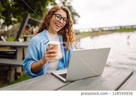 Portrait of a young woman in a blue shirt with a laptop at a table in an outdoor cafe near a lake. Portrait of a young woman in a blue shirt with a laptop at a table in an outdoor cafe near a lake. 126593856