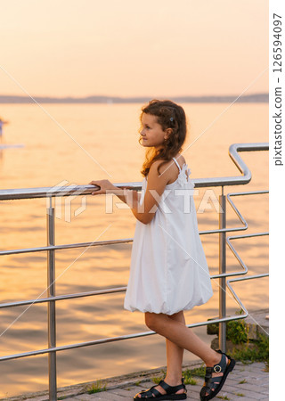 Child girl stands on pier overlooking body of water. She is wearing white dress and is smiling 126594097