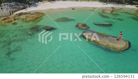 A woman enjoys a peaceful moment standing on a rock surrounded by clear turquoise water and sandy beach. Lush greenery frames the stunning coastal landscape, creating an idyllic setting. A woman enjoys a peaceful moment standing on a rock surrounded by clear turquoise water and sandy beach. Lush greenery frames the stunning coastal landscape, creating an idyllic setting. 126594131