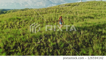 Young woman walking across lush green hills, relishing the vastness of nature and embracing the freedom of open space beneath a dramatic, cloudy sky 126594142