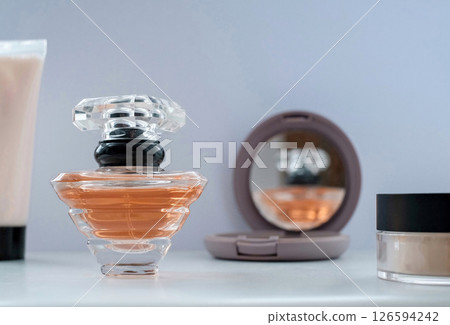 Elegant flat lay of a perfume bottle with peach-colored fragrance, compact mirror reflecting the bottle, and makeup items on a white surface.  126594242
