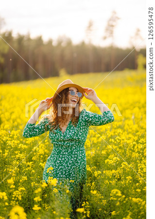 Beautiful woman in bright dress and elegant hat walks and has fun in rapeseed field. Nature, rest. 126595478