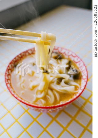 person is eating noodles with chopsticks. colorful bright red plate of ramen sits on yellow table. person is eating noodles with chopsticks. colorful bright red plate of ramen sits on yellow table. 126595522