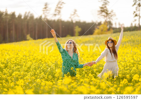 Two young women in beautiful dresses walk in a field with yellow blooming rapeseed. Concept of fun. 126595597