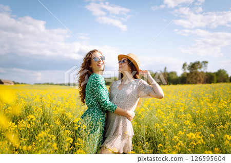 Two young women in beautiful dresses walk in a field with yellow blooming rapeseed. Concept of fun. Two young women in beautiful dresses walk in a field with yellow blooming rapeseed. Concept of fun. 126595604