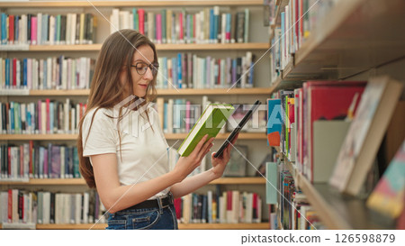 A young student in a university library selects a book from a shelf while holding a tablet and book, traditional and digital research methods in an academic setting, surrounded by bookshelves.  126598879
