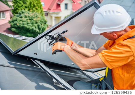Worker building solar panel system on metal rooftop of house. Man technician preparing to install photovoltaic solar module outdoors. Alternative, green and renewable energy generation concept. 126599426