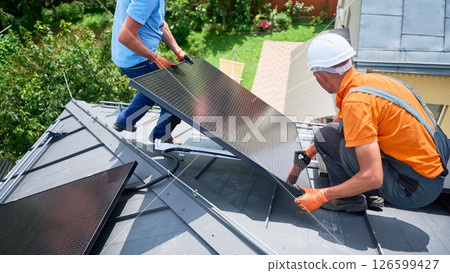Workers building solar panel system on rooftop of house. Two men installers in helmets installing photovoltaic solar module outdoors. Alternative, green and renewable energy generation concept. Workers building solar panel system on rooftop of house. Two men installers in helmets installing photovoltaic solar module outdoors. Alternative, green and renewable energy generation concept. 126599427