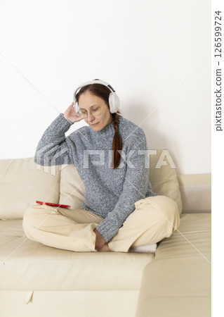 Woman wearing headphones listening to music on smartphone while relaxing on sofa Woman wearing headphones listening to music on smartphone while relaxing on sofa 126599724