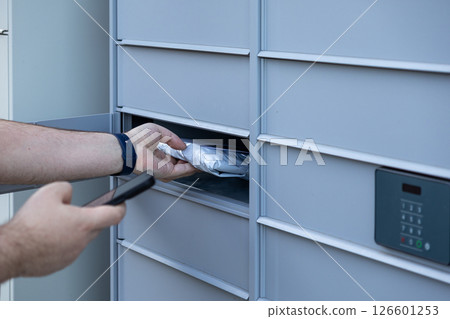 Customer hand with smartphone retrieving package from automated parcel locker using keypad for modern self-service delivery access Customer hand with smartphone retrieving package from automated parcel locker using keypad for modern self-service delivery access 126601253