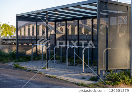 Empty urban bike shelter with metal racks and glass panels reflecting city bridge at sunset for sustainable transport concepts Empty urban bike shelter with metal racks and glass panels reflecting city bridge at sunset for sustainable transport concepts 126601279