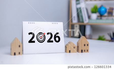 Desk calendar showing year 2026 with a dartboard target replacing the zero, Surrounded by small wooden houses symbolizing goal setting, planning, and future investment in real estate. Desk calendar showing year 2026 with a dartboard target replacing the zero, Surrounded by small wooden houses symbolizing goal setting, planning, and future investment in real estate. 126601334