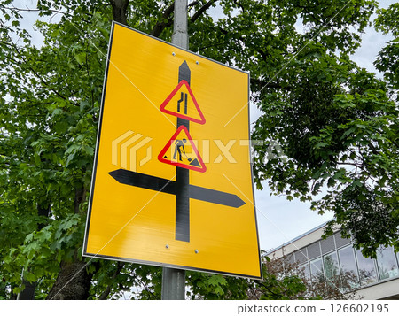 Bright yellow temporary road sign displays merging lanes, road work symbols, and directional arrows indicating traffic changes and providing detour guidance near construction zone. Bright yellow temporary road sign displays merging lanes, road work symbols, and directional arrows indicating traffic changes and providing detour guidance near construction zone. 126602195