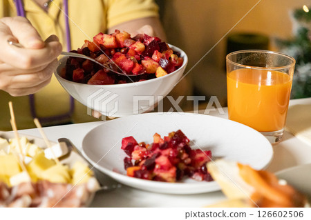 person serving beet vinaigrette salad with vegetables and peas from a white bowl onto a plate, with fresh orange juice on the table 126602506