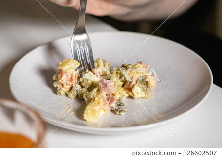 person eating Olivier salad with fork on a plate, close-up of ingredients like potatoes, ham, pickles, and carrots 126602507