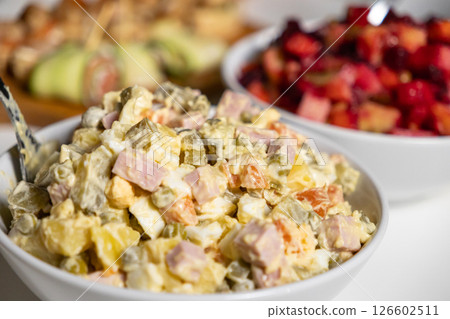 classic olivier salad with potatoes, ham, pickles, eggs, and mayonnaise in a white bowl, with vinaigrette salad in the background on a festive table 126602511